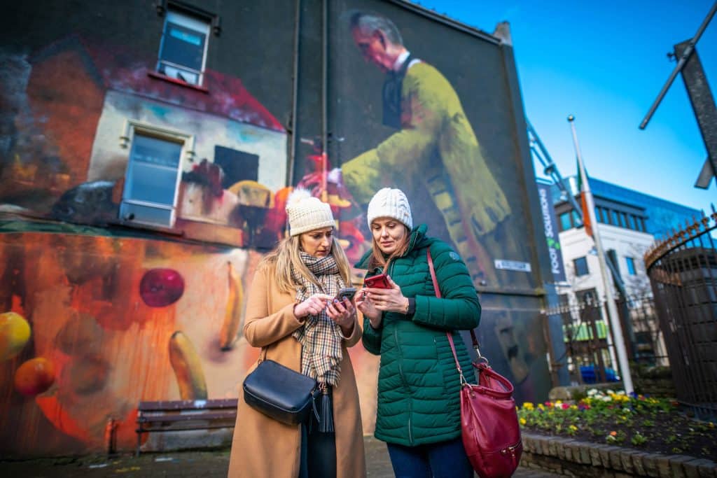 Two women in winter clothes stand on a city street looking at a mobile phone together, with a colourful mural featuring a large figure and fruit painted on the building wall behind them.