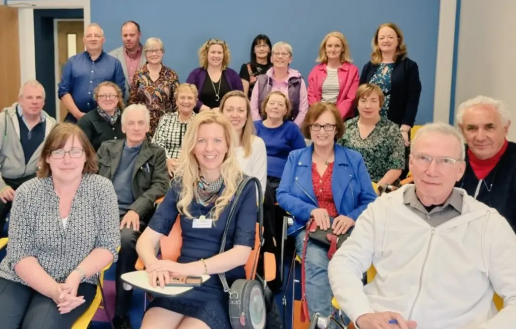 A group of 20 adults, men and women, are sitting and standing in a classroom setting, smiling at the camera. The room has blue walls and chairs arranged in rows.