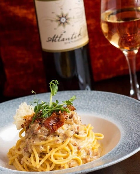 A plate of creamy pasta topped with herbs and crispy bits, served with a glass of white wine and a bottle labelled "Atlantik" in the background. The setting is elegant and inviting.