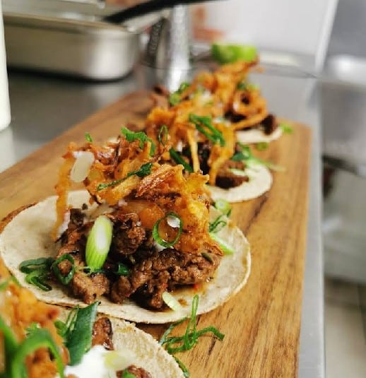 Three tacos on a wooden board, topped with seasoned meat, crispy fried onions, chopped spring onions, and a creamy sauce. The background shows a blurred kitchen setting.