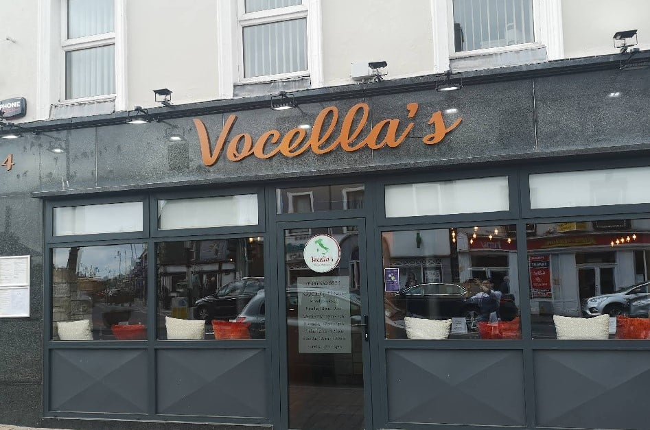 Street view of "Vocella's" restaurant with black and grey exterior, large front windows, and orange sign above the entrance. Reflections and parked cars can be seen through the windows.