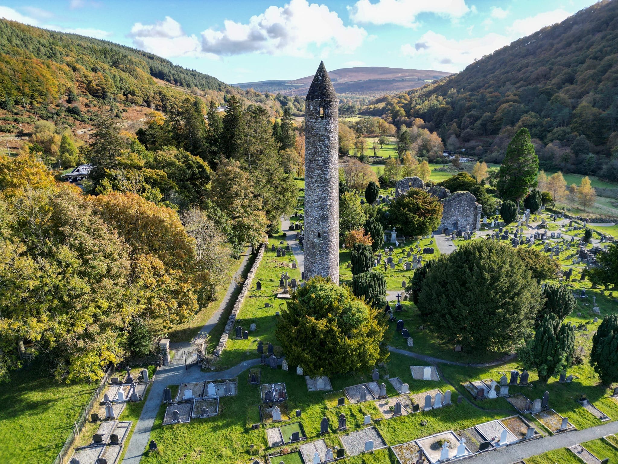 A tall, round stone tower stands in the centre of a historic cemetery with gravestones, surrounded by lush trees and rolling hills under a bright blue sky with scattered clouds.