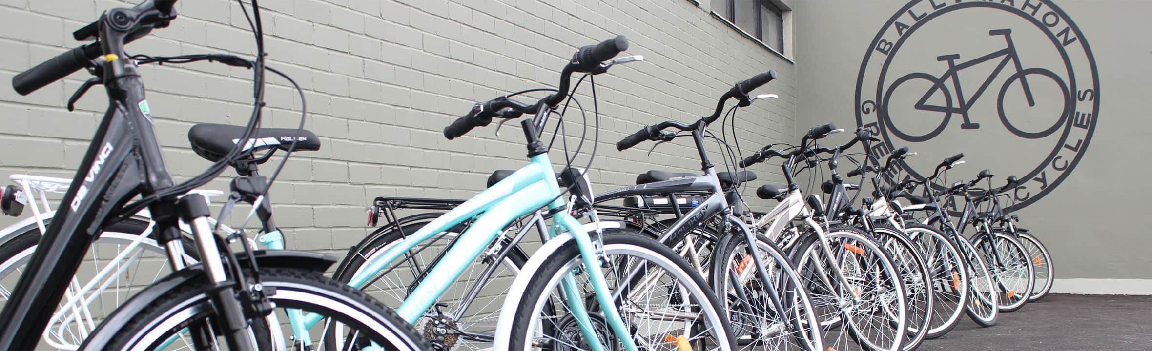 A row of bicycles is parked along a grey brick wall outside a building with a large circular bike shop logo painted on the wall.