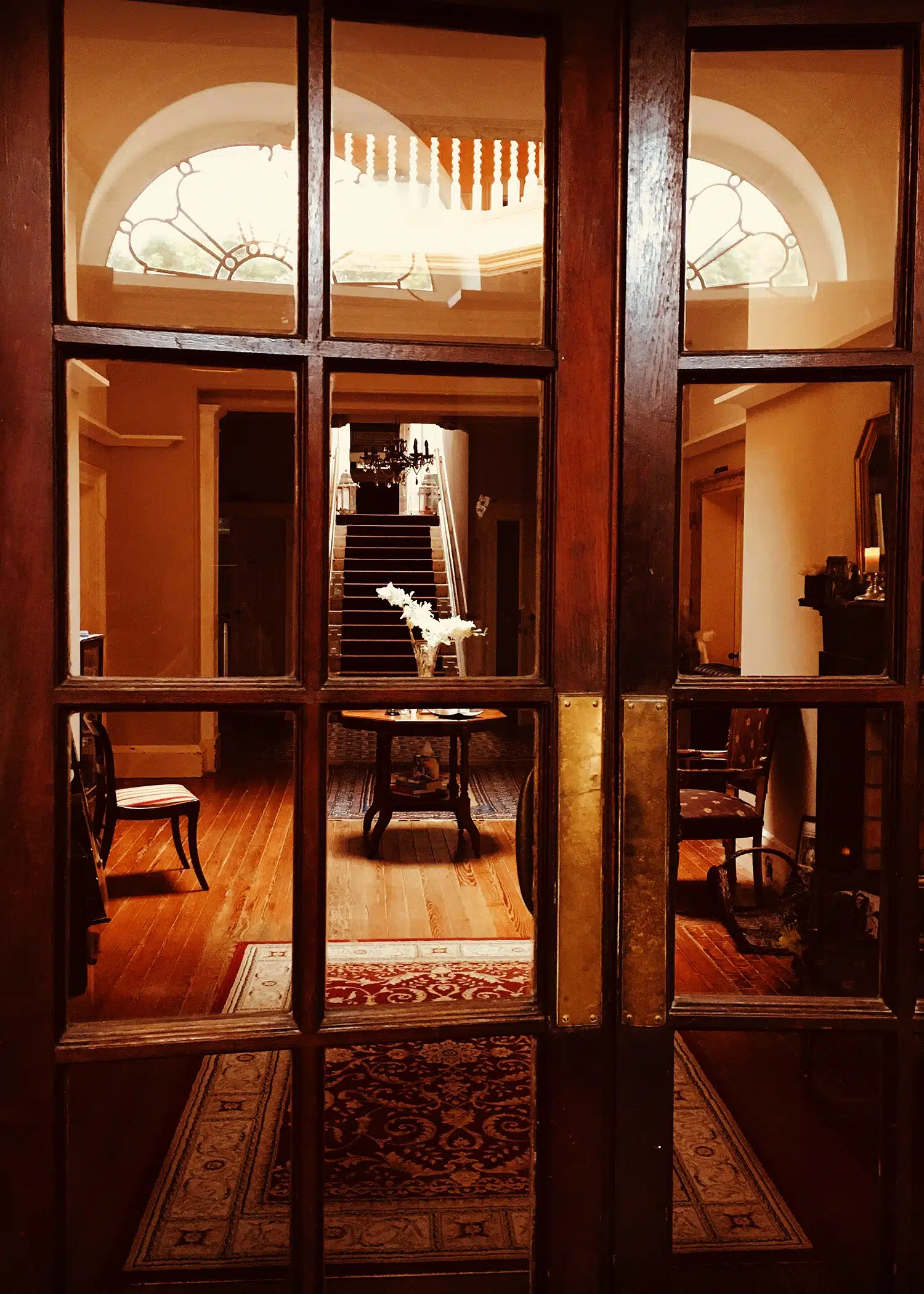 View through wooden glass doors into a warmly lit hall with a round table holding a vase of white flowers, polished wooden floors, chairs, ornate rugs, and a staircase in the background.