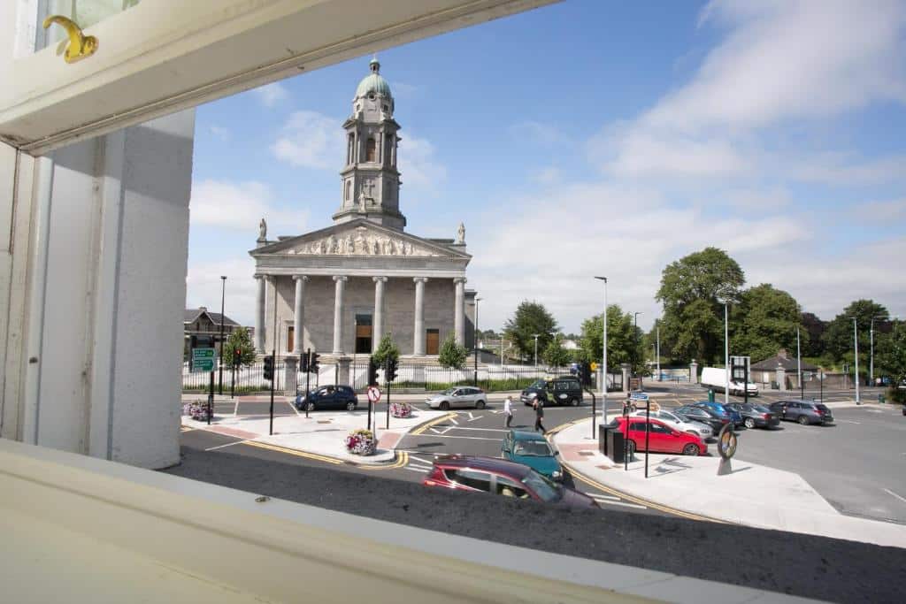 View from a window overlooking a roundabout with several parked and moving cars, and a large historic stone building with columns and a clock tower in the background, under a partly cloudy sky.