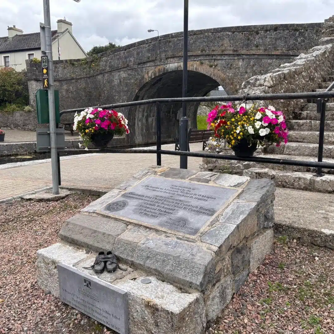 A stone memorial with plaques and small shoes sits in front of a stone bridge, with colourful flower baskets on railings and houses in the background. The sky is cloudy.