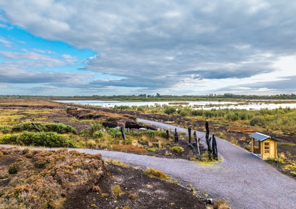 A gravel path curves through a landscape of peat bogs and scrub vegetation under a mostly cloudy sky, with a small wooden shelter and several upright charred posts beside the path.