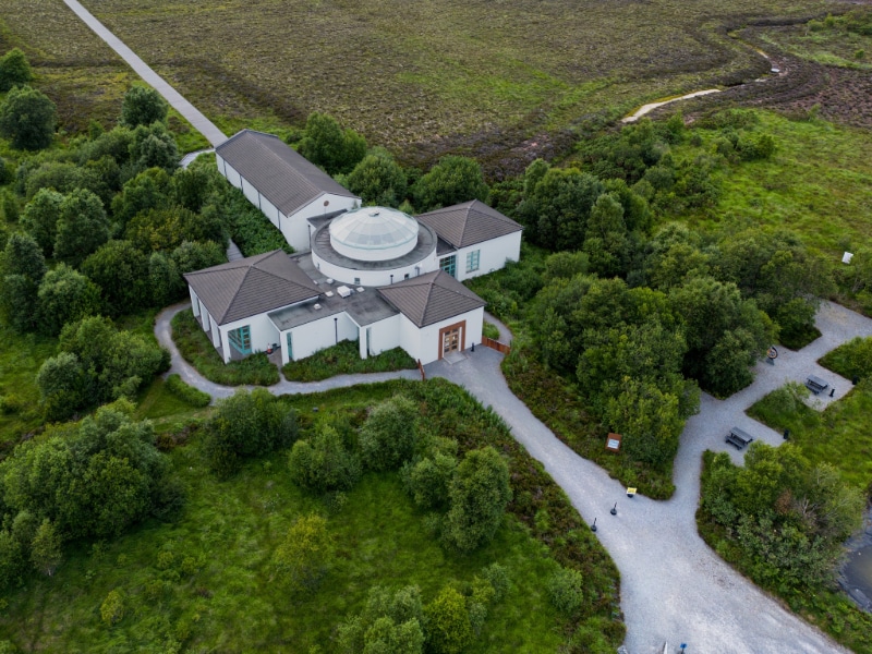 Aerial view of a white, modern building with a central glass dome surrounded by lush green trees and winding gravel paths in a rural, grassy landscape.