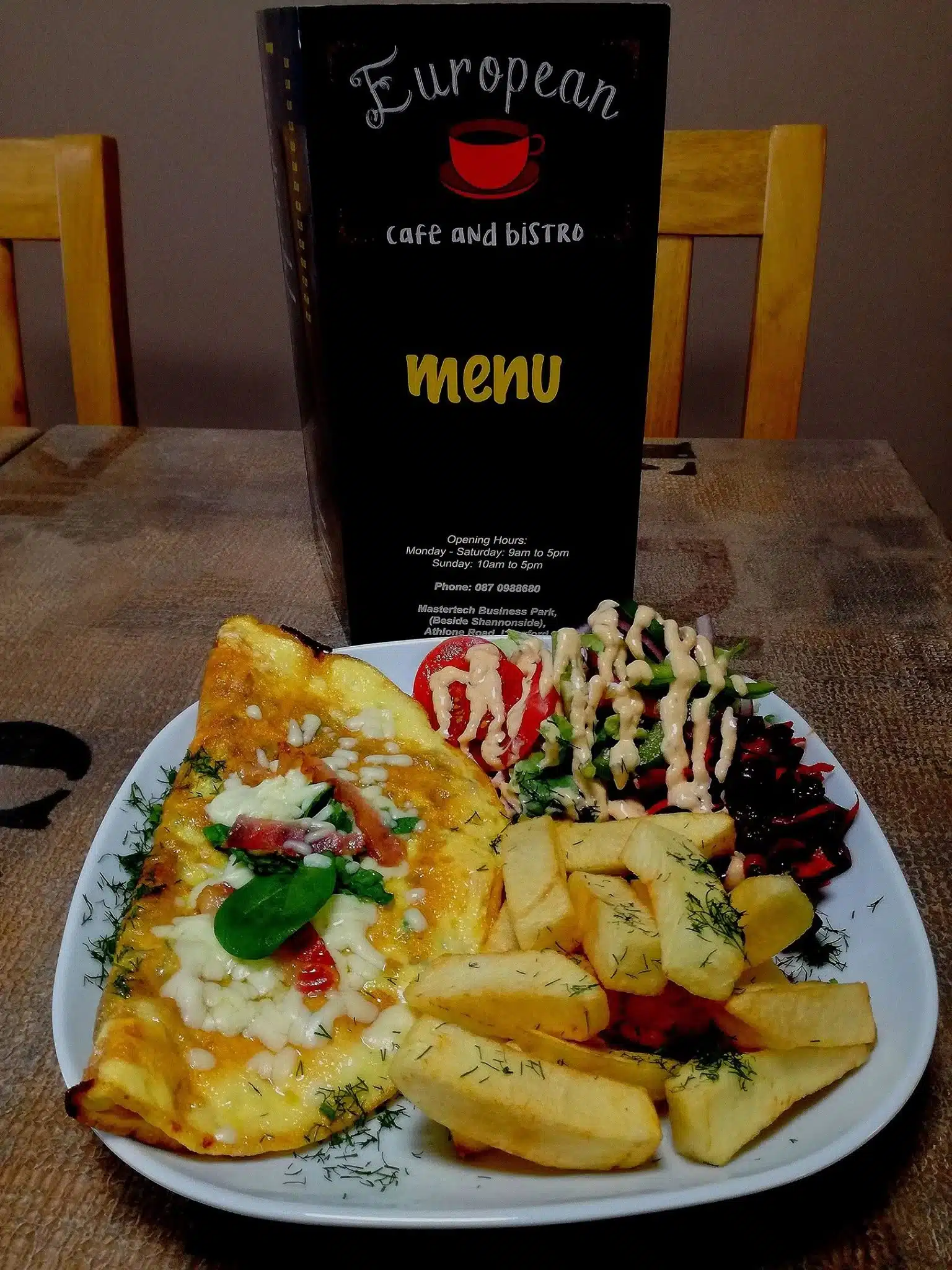 A plate with an omelette, chips, salad with dressing, and beetroot slices sits on a table. A standing menu from "European Cafe and Bistro" is in the background, with two wooden chairs visible.