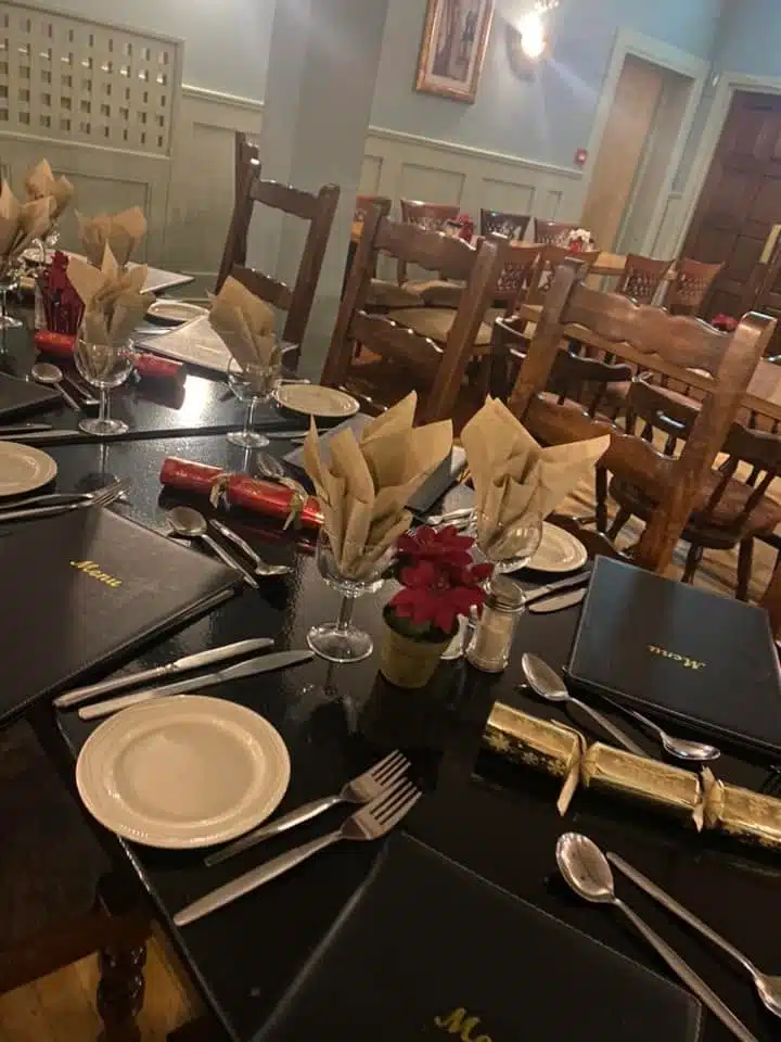 A dining table set for a meal with menus, plates, cutlery, wine glasses, crackers, and serviettes folded in glasses. Wooden chairs surround the table in a warmly lit room with light-coloured walls.