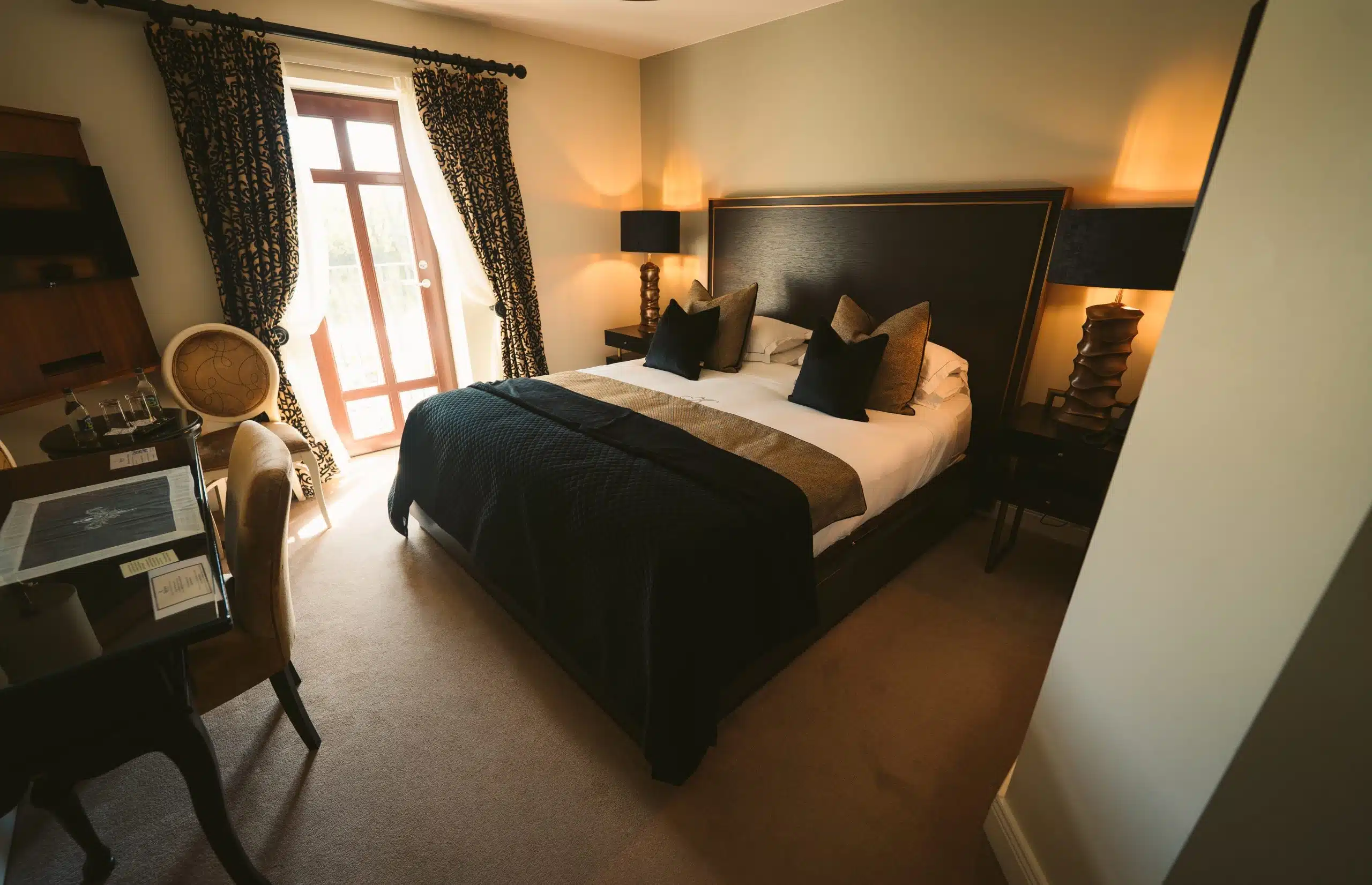 A modern, neatly arranged hotel room with a large bed, dark pillows and duvet, two bedside tables with lamps, a desk with a chair, and a window with patterned curtains letting in natural light.