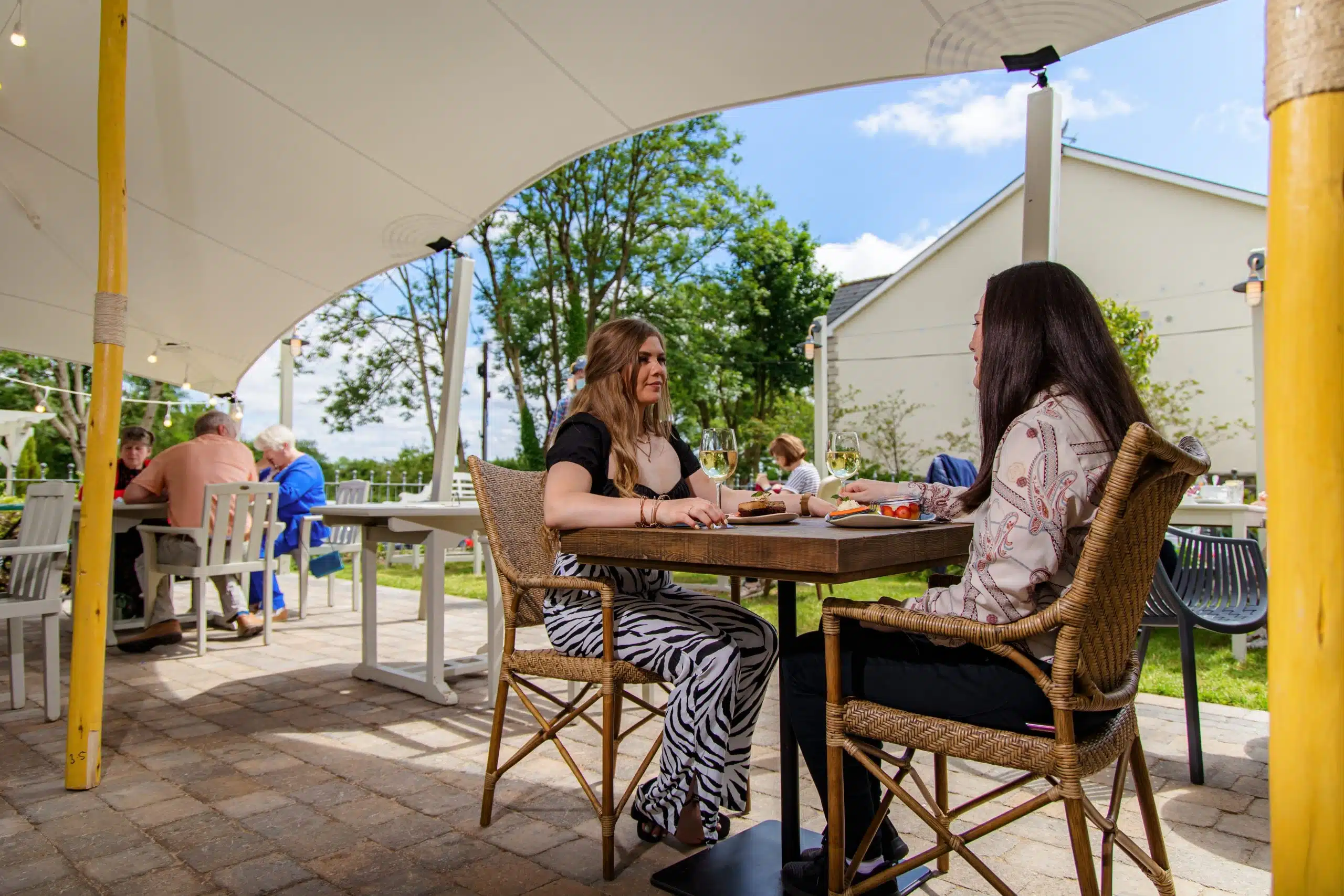 Two women sit at an outdoor restaurant table under a white canopy, eating and talking. Other diners are seated at nearby tables. Trees and a building are visible in the background on a sunny day.