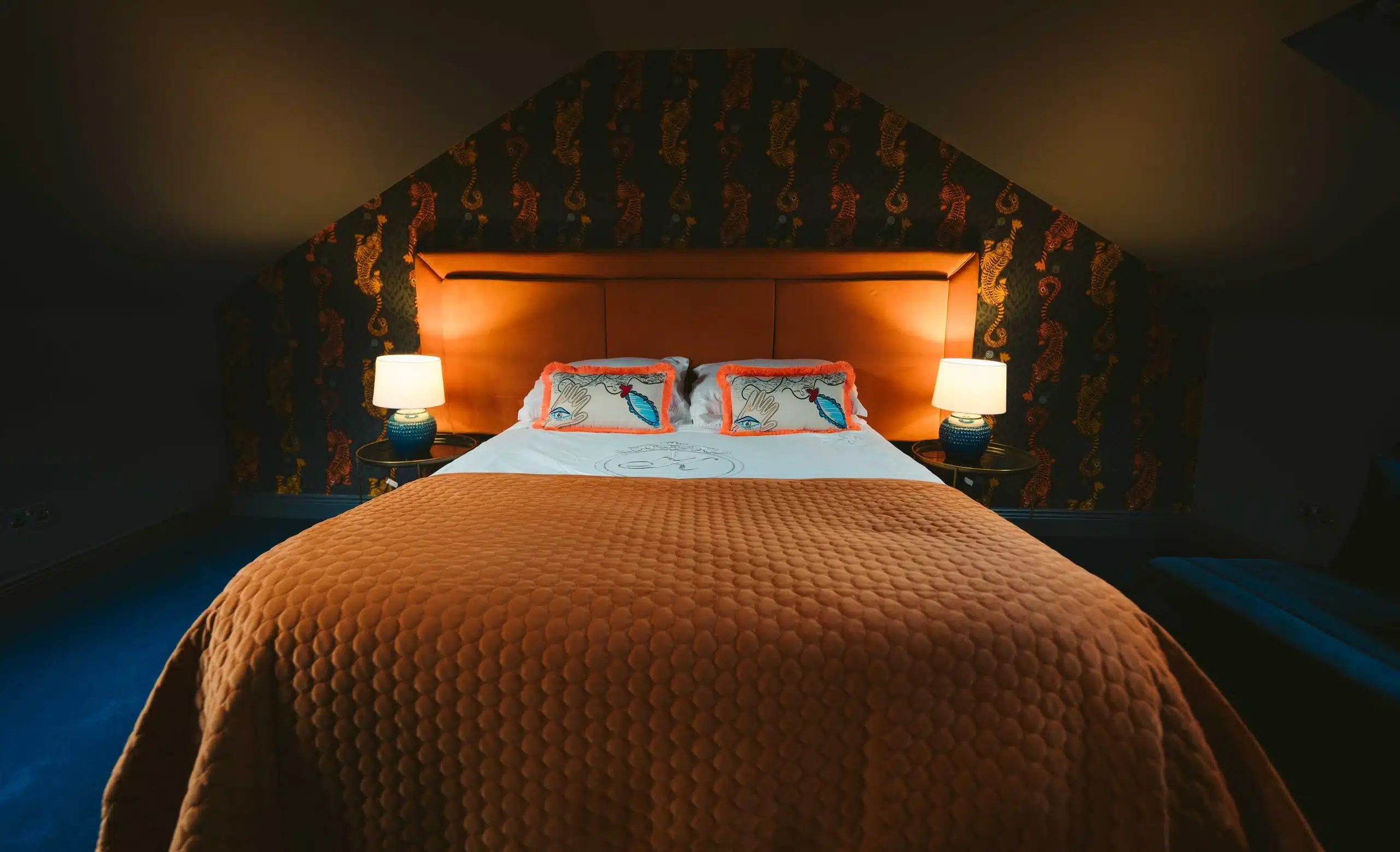 A cosy bedroom with a large orange quilted bed, two pillows featuring bird designs, two bedside tables with lamps, and a dark wallpaper with a seahorse pattern.