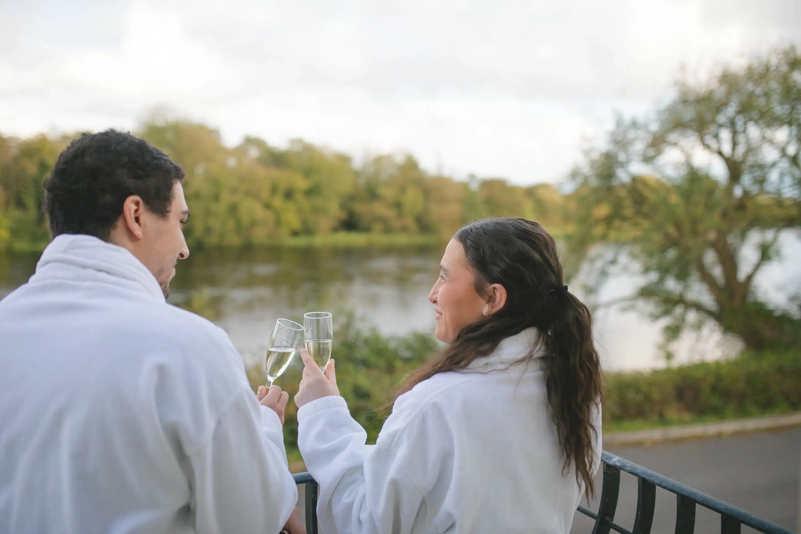 Two people in white dressing gowns stand on a balcony by a lake, holding and clinking champagne glasses, with trees and water in the background.