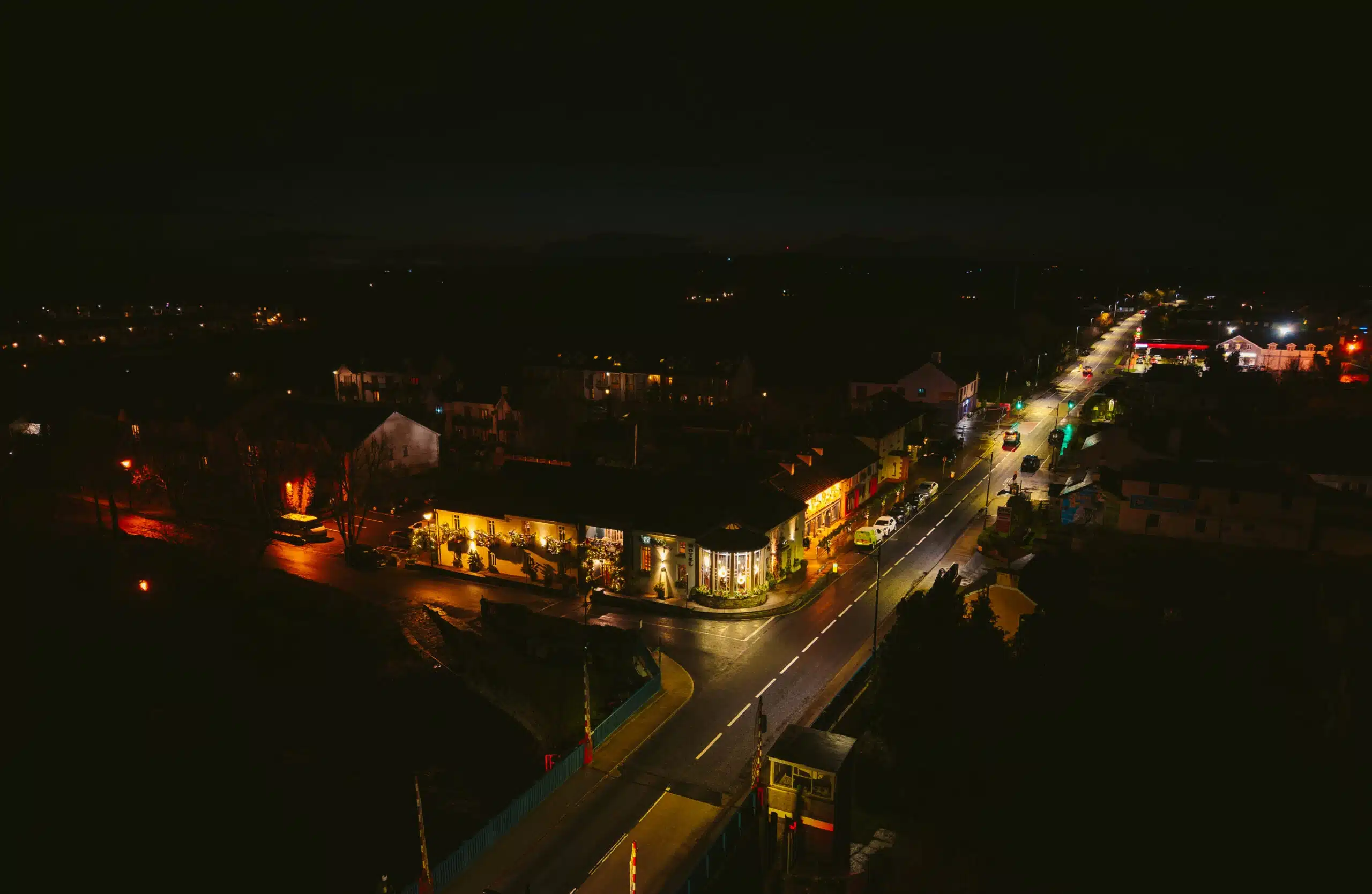 A night-time aerial view of a small town with warmly lit buildings and streetlights lining the main road, creating a cosy, inviting atmosphere against the dark surroundings. Cars are parked along the streets and some are driving by.