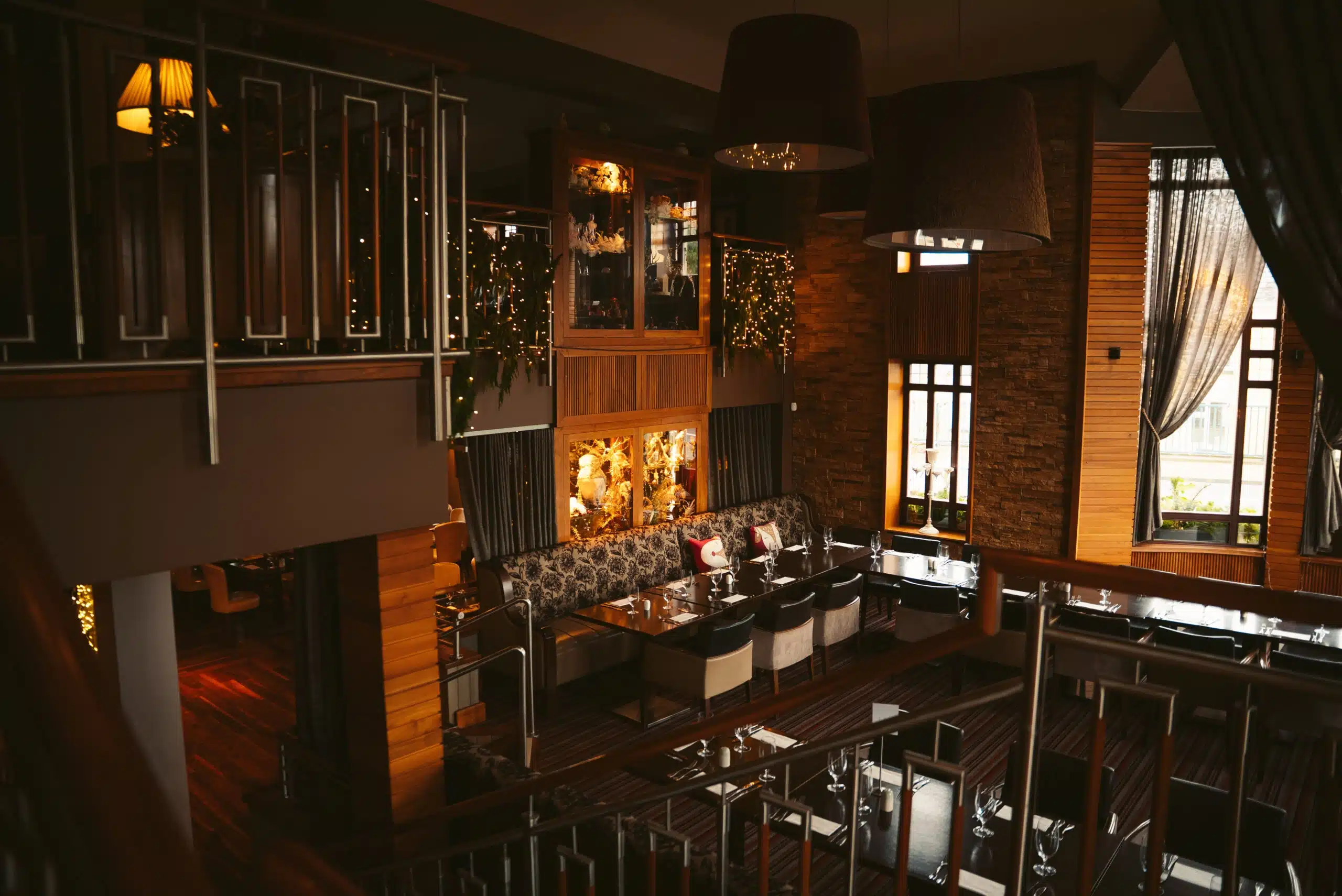 A cosy, dimly lit restaurant interior with wooden accents, fairy lights, large windows, cushioned bench seating, and elegant tables set for dining. Warm lighting creates an inviting and intimate atmosphere.