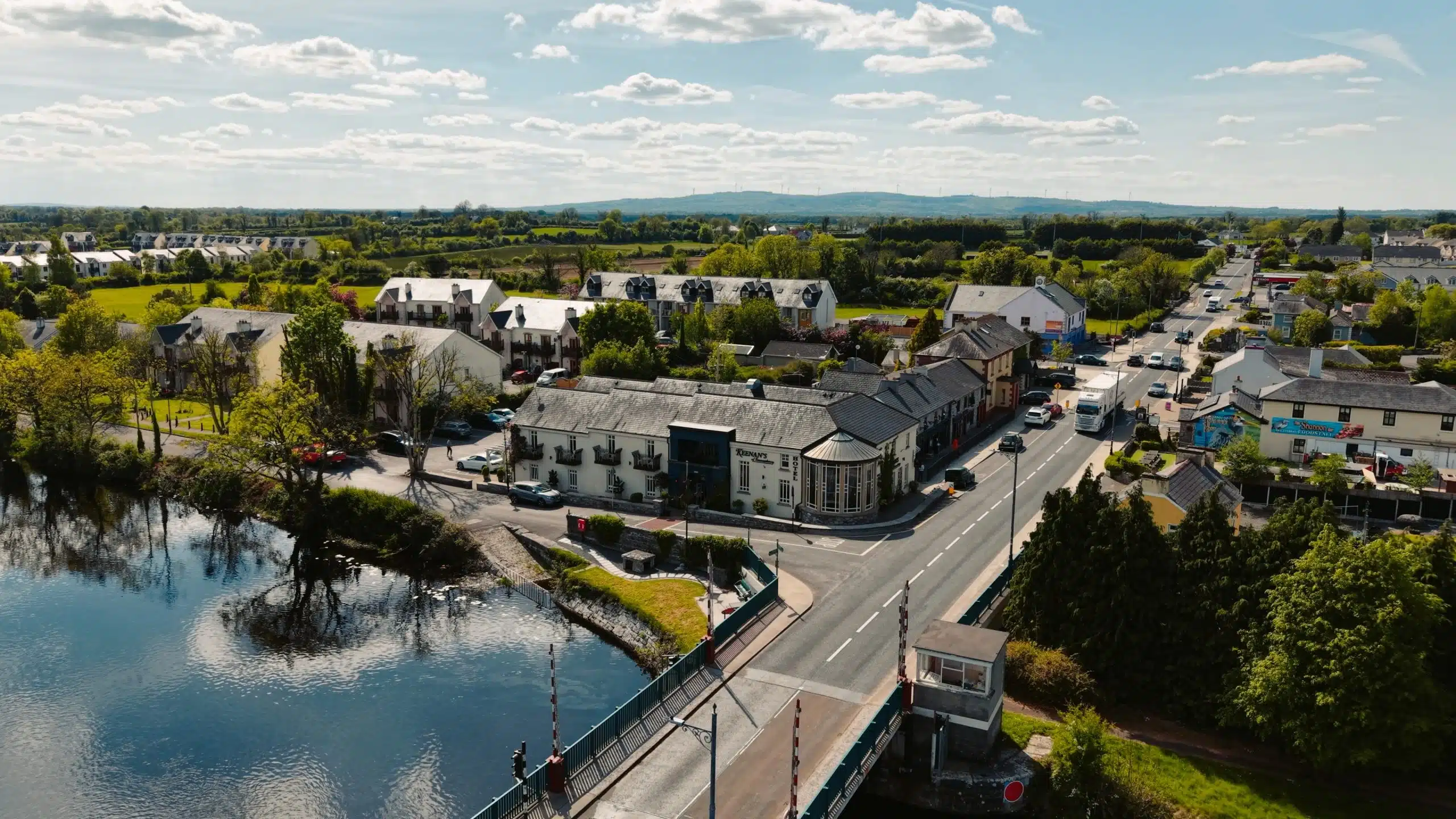 Aerial view of a small town with a bridge crossing over a river, surrounded by houses, trees, roads, and cars under a blue sky with scattered clouds.
