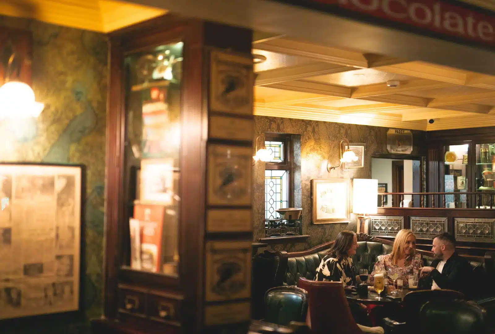 Three people sit at a table in a cosy, dimly lit pub with vintage decor, talking and enjoying drinks. The warm lighting and wooden furnishings create a welcoming, relaxed atmosphere.