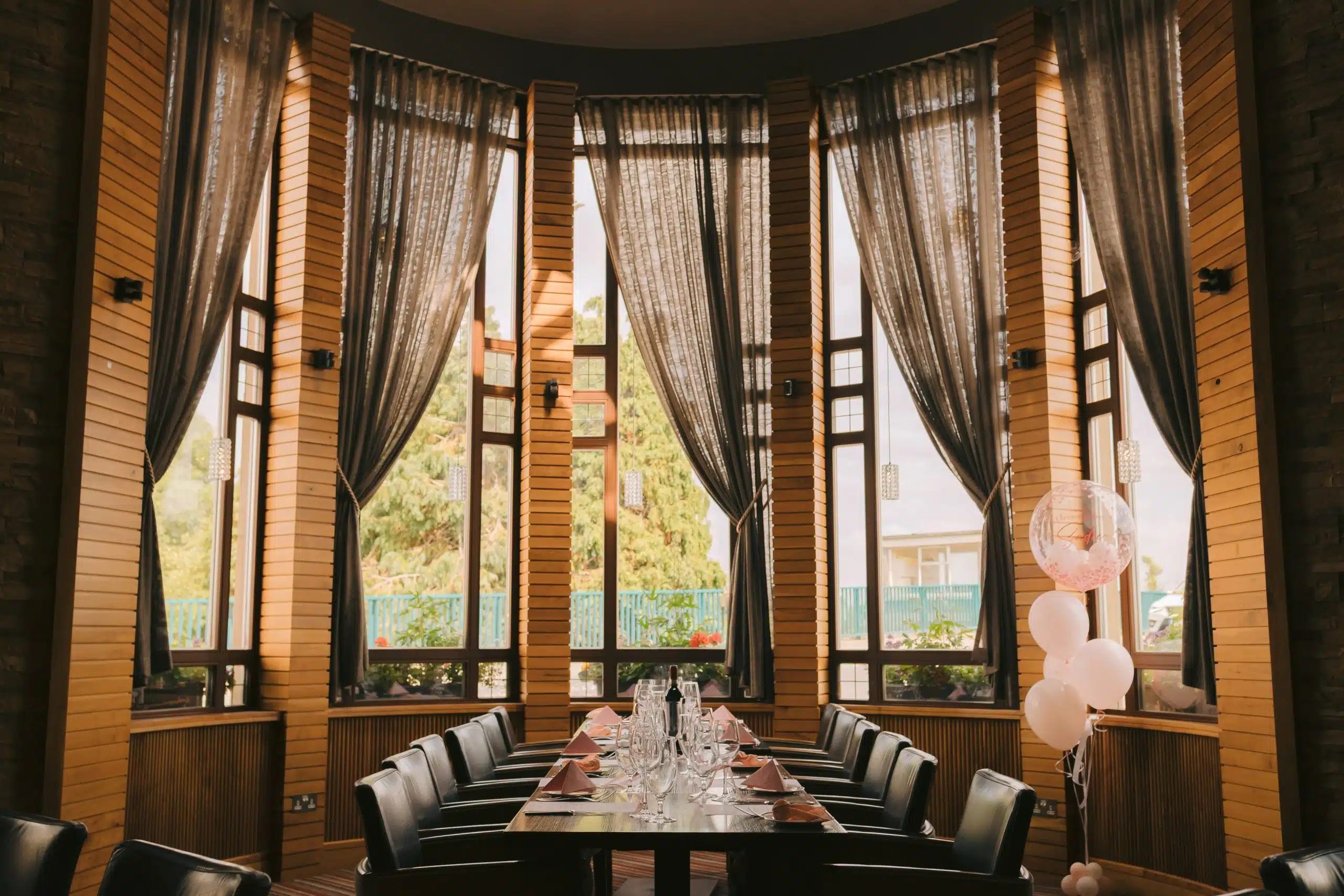 A long dining table set with glassware and serviettes is surrounded by black chairs in a restaurant with tall windows and sheer curtains. A bunch of white and pink balloons decorates one end of the table.