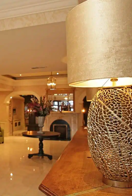 A close-up of a decorative gold lamp on a wooden table in a warmly lit, elegant hotel lobby with marble floors, a round table with flowers, arched doorways, and a fireplace in the background.