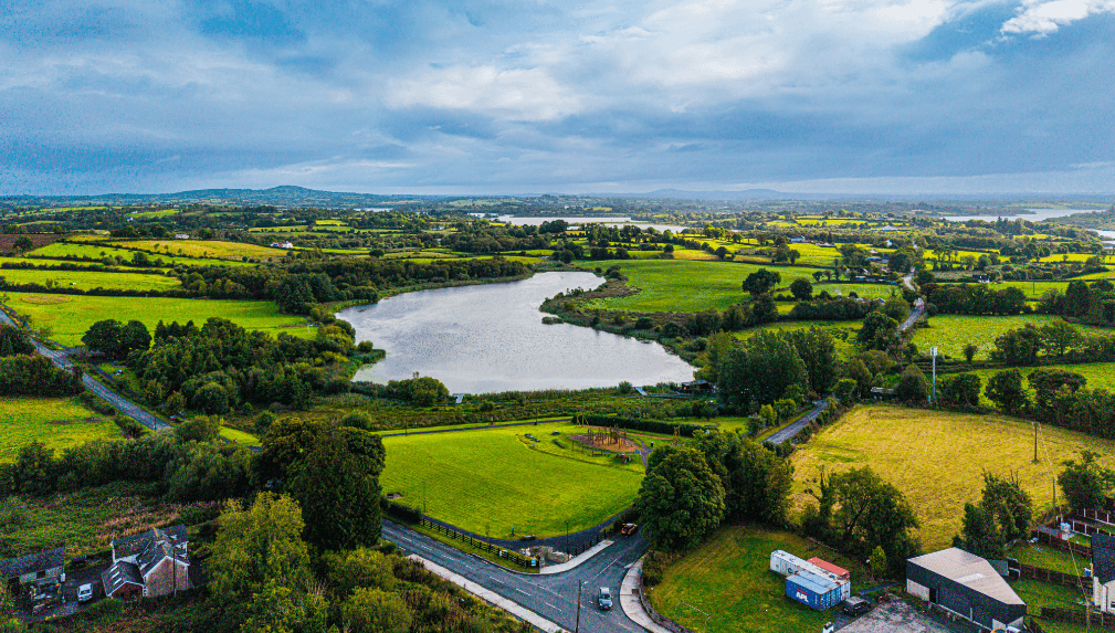 Aerial view of a lush, green countryside with patchwork fields, a large pond or lake in the centre, scattered houses, trees, and roads under a cloudy sky.