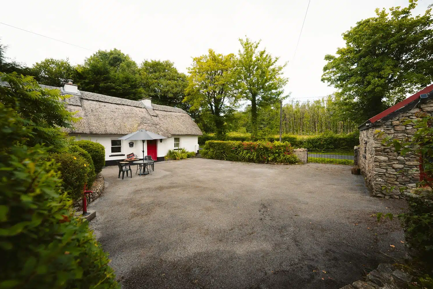 A quaint white cottage with a thatched roof and red door is surrounded by lush greenery. An outdoor table with chairs and a parasol sits on a gravel courtyard beside a stone wall and leafy trees.
