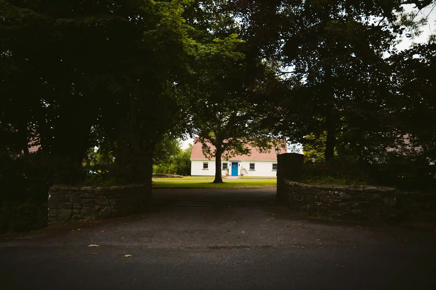 A white house with a red roof and blue door stands beyond a stone wall and tree-lined drive, surrounded by greenery and shaded by large trees.
