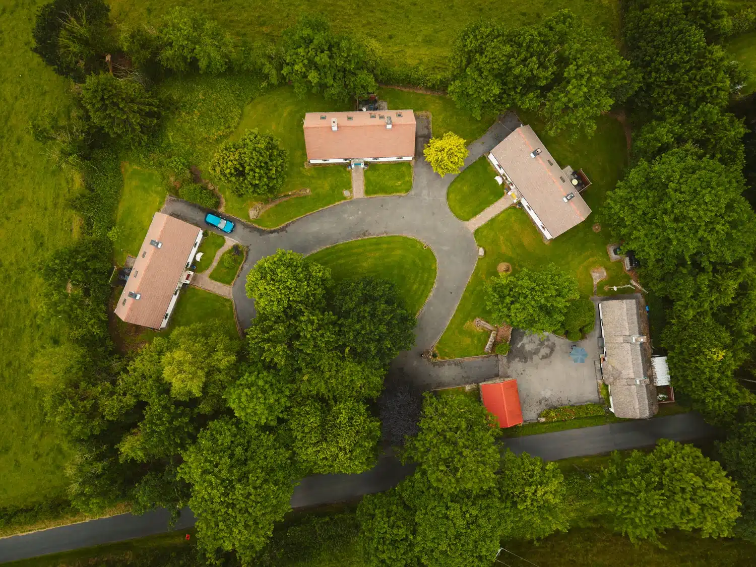 Aerial view of three rectangular houses with brown roofs arranged around a circular drive, surrounded by green trees and grass, with a road running along the bottom of the image.