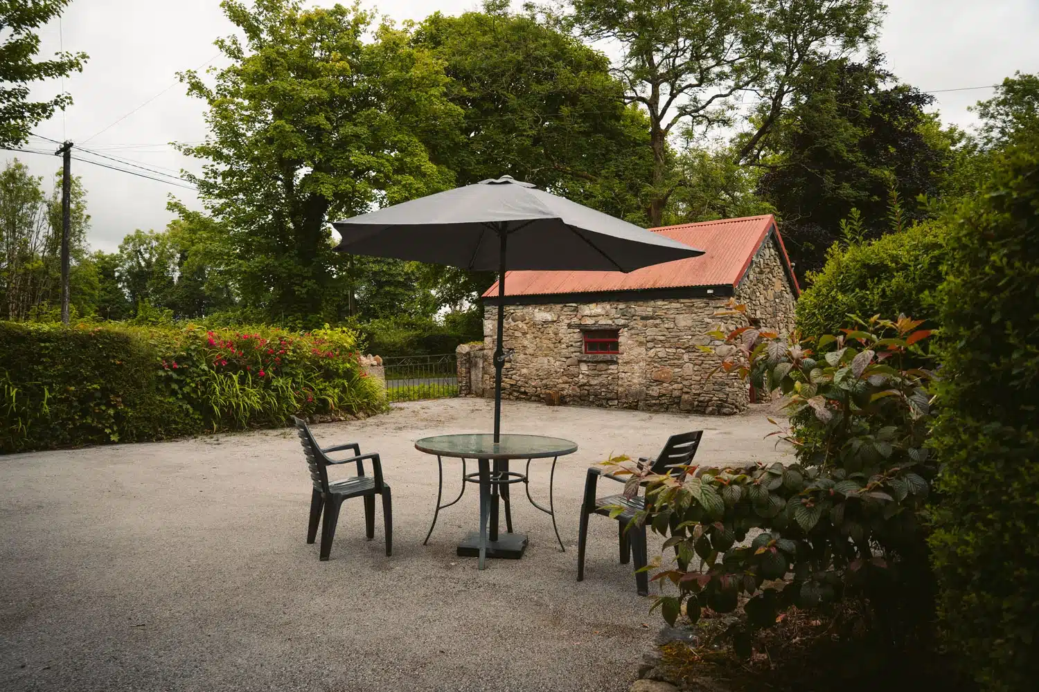 A round glass patio table with four black chairs and a large black umbrella sits on a paved area, with a stone building, greenery, and trees in the background.