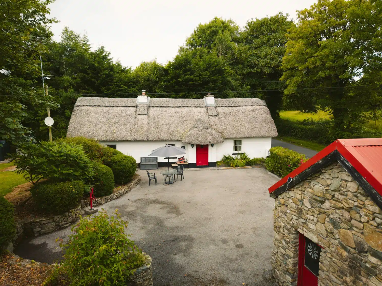 A white thatched-roof cottage with a bright red door sits surrounded by trees and greenery. There is a stone outbuilding with a red roof in the foreground and garden furniture in the courtyard.