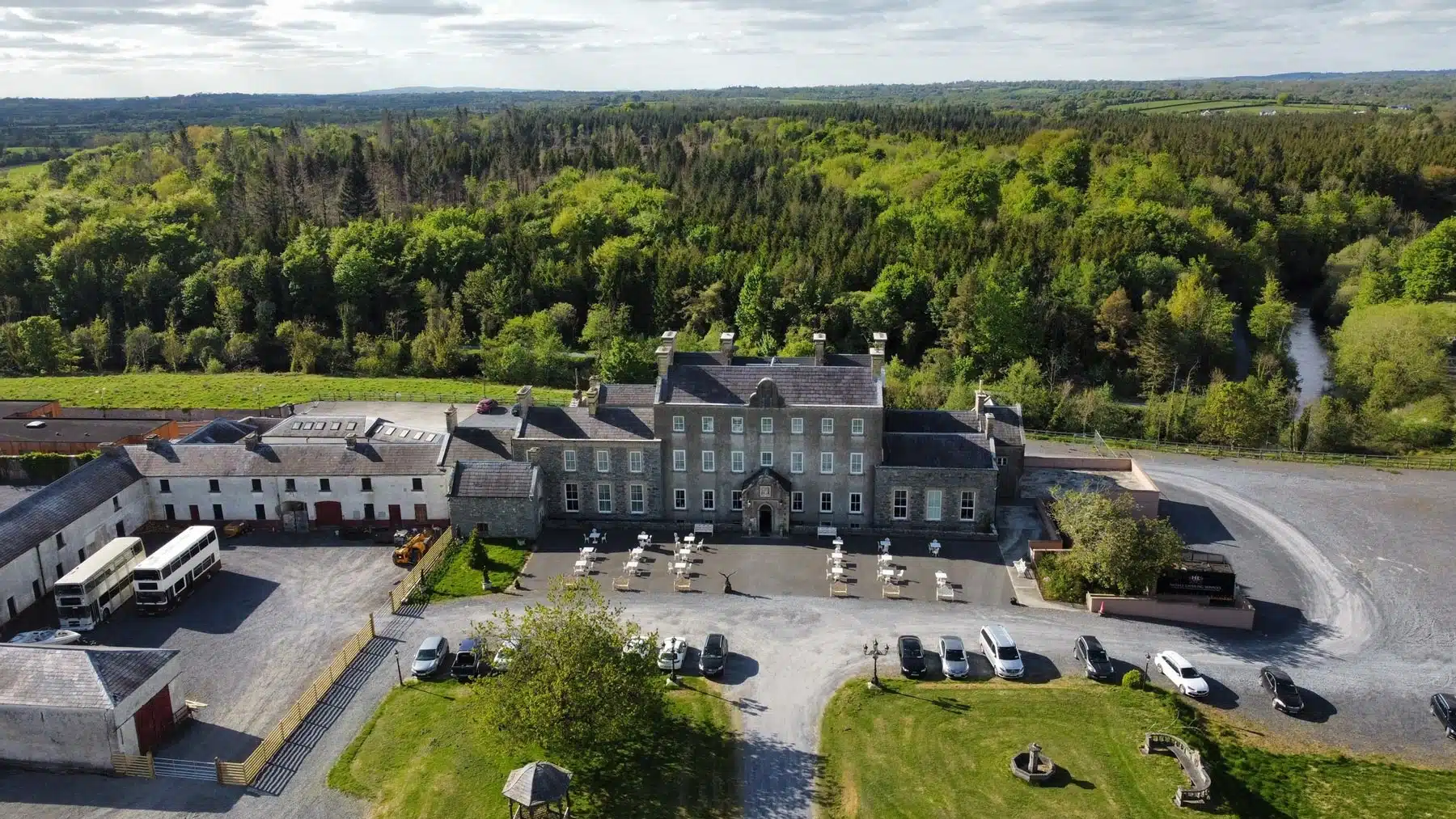 Aerial view of a large historic building with a courtyard, outdoor seating, parked cars, and surrounded by lush green trees and countryside under a partly cloudy sky.