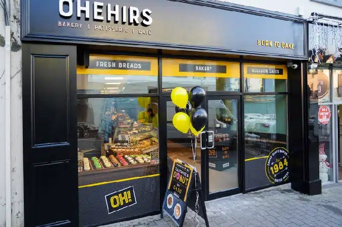 Exterior of OHEHIRS bakery and café, with black and yellow signage, window displays of baked goods, a cluster of black and yellow balloons, and a sign advertising doughnuts on the pavement.