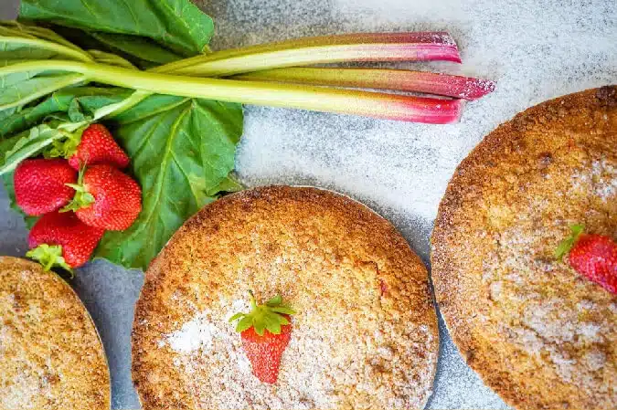 Three golden pies topped with sugar and a single strawberry sit on a light surface next to fresh rhubarb stalks and strawberries with green leaves.