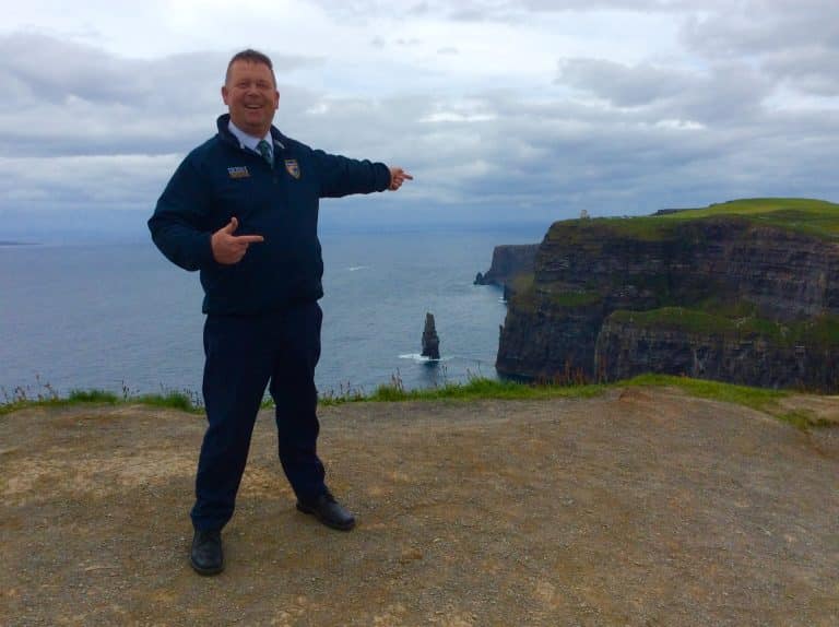 A smiling man in a dark jacket stands on a cliffside path, pointing towards the Cliffs of Moher and a sea stack rising from the ocean below under a cloudy sky.
