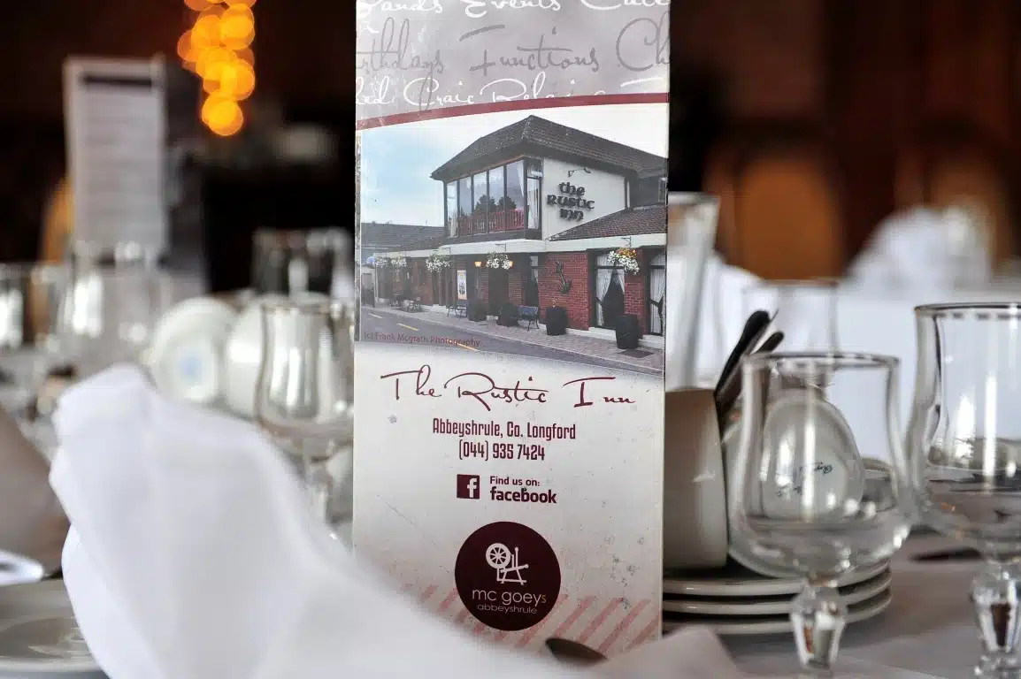 A close-up of a restaurant menu card for "The Rustic Inn" on a formal table set with white linen, glassware, and cutlery. The card displays the inn’s photo, contact details, and social media icons.