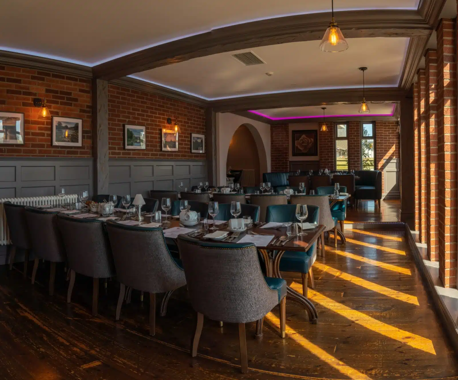 A stylish restaurant dining room with wooden floors, brick walls, elegant dark chairs, set tables with glassware, and warm sunlight streaming through large windows. Framed art decorates the walls.