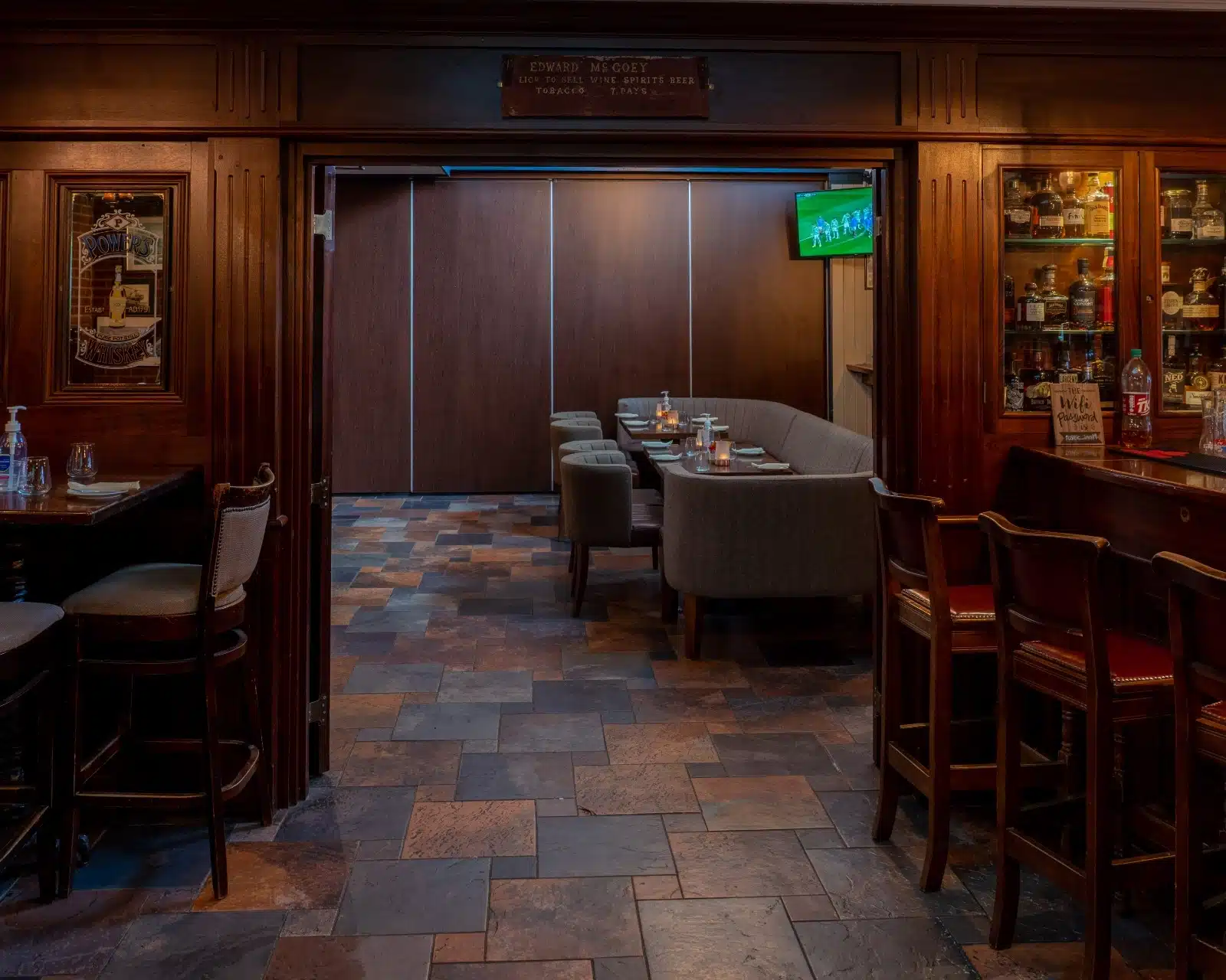 A cosy pub interior with wooden décor, padded chairs, and a table set for dining. A TV on the wall displays a sports match. The room has dim lighting, tiled floor, and bottles visible in a nearby glass cabinet.
