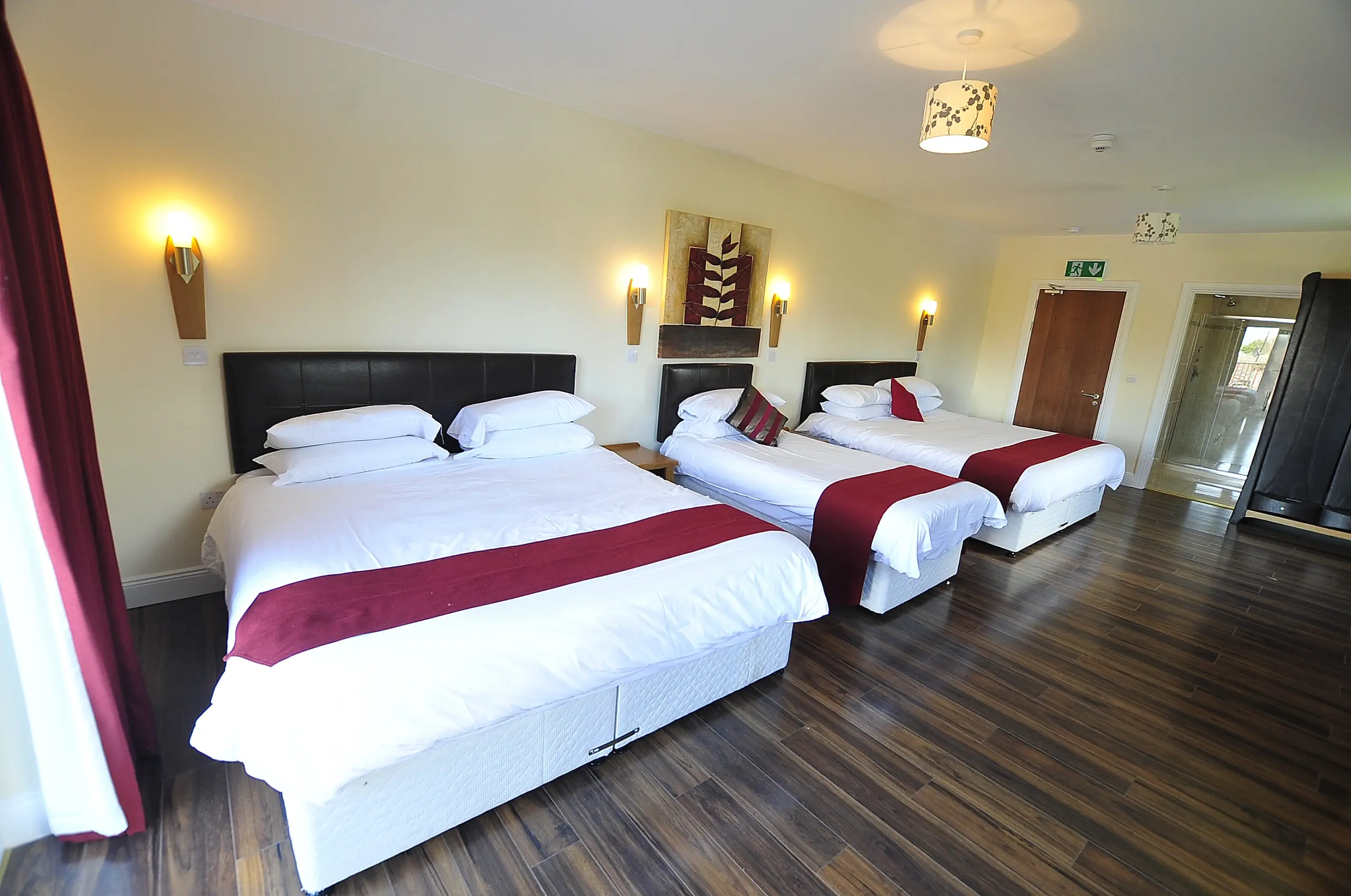 A hotel room with three double beds, each with white bedding and a maroon runner, wall-mounted lights above the beds, wooden flooring, modern decor, and a door leading to another area.