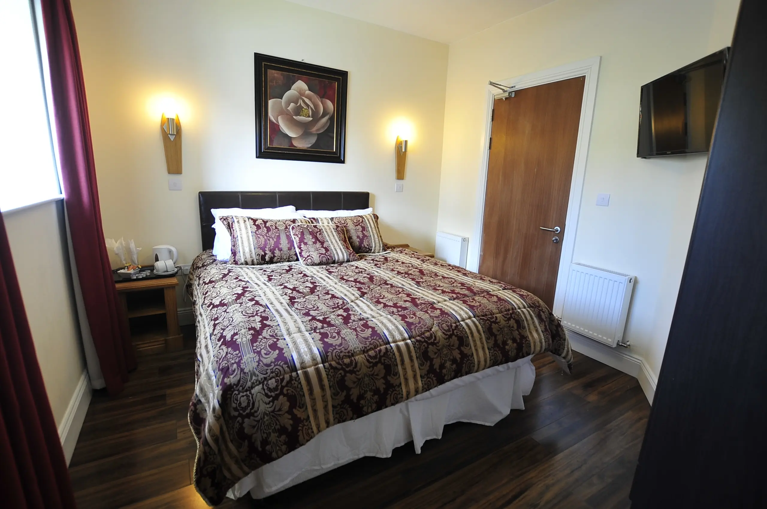 A neatly made double bed with a patterned burgundy and gold duvet in a cosy bedroom, featuring wall lamps, a flower painting, wooden floor, a small bedside table with a kettle, a door, and a wall-mounted TV.