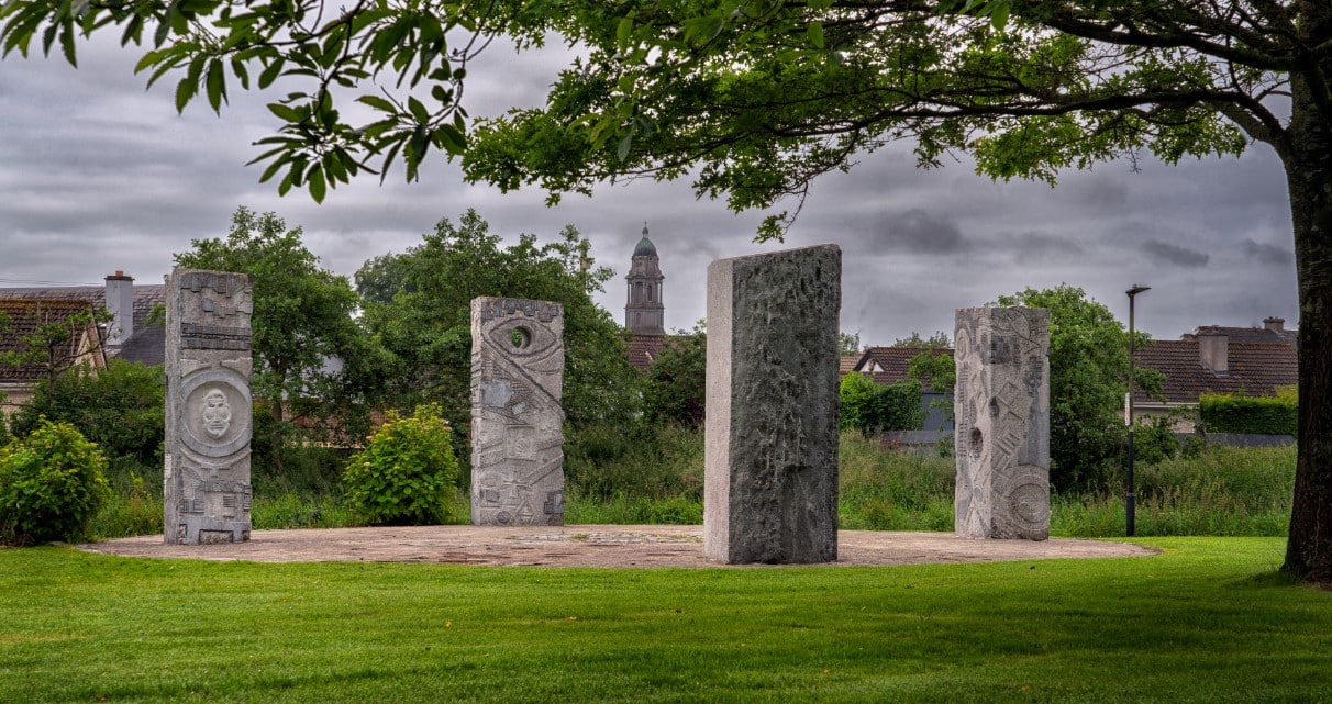 Five large carved stone pillars stand in a circle on a grassy area under a tree, with houses and a church tower visible in the background on a cloudy day.