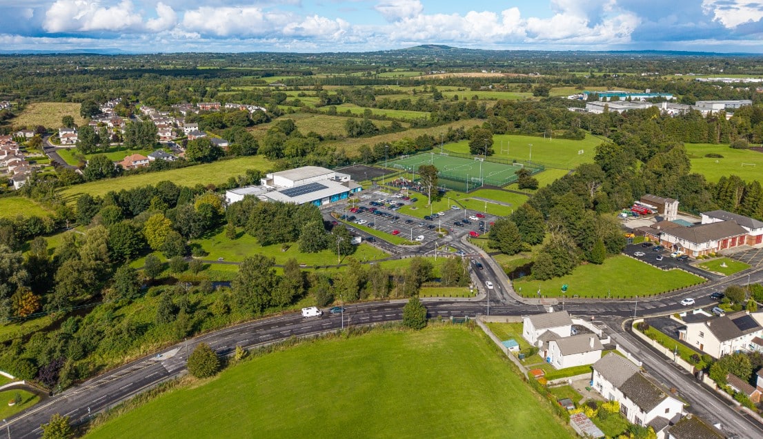 Aerial view of a suburban area with green fields, trees, sports courts, a large car park, and residential houses, all under a partly cloudy sky with distant hills in the background.