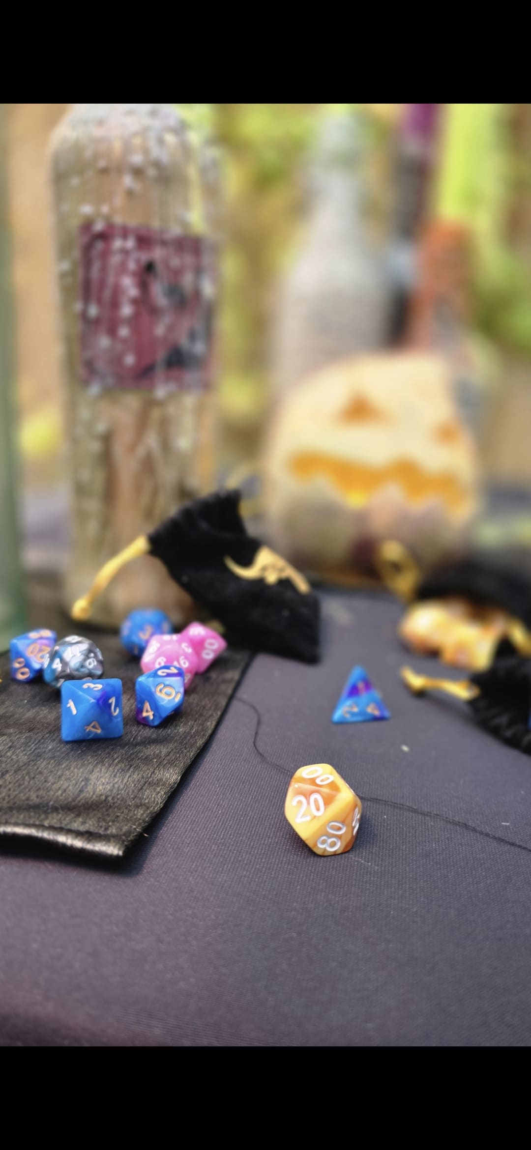 A set of colourful polyhedral dice, including an orange 20-sided die, are scattered on a black cloth near velvet bags, bottles, and a carved pumpkin in the background.