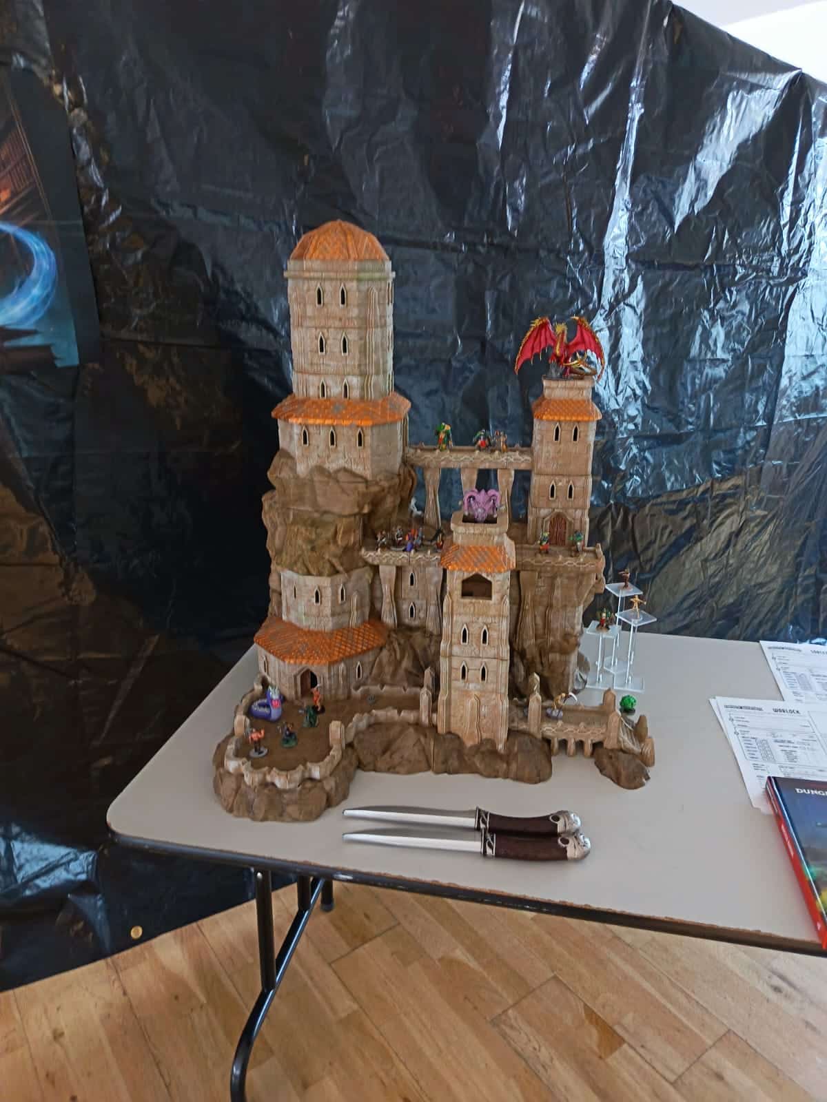 A detailed tabletop castle model with towers, figurines, and a red dragon on top is displayed on a table. Two ornate daggers are placed in front, along with papers and books to the side.