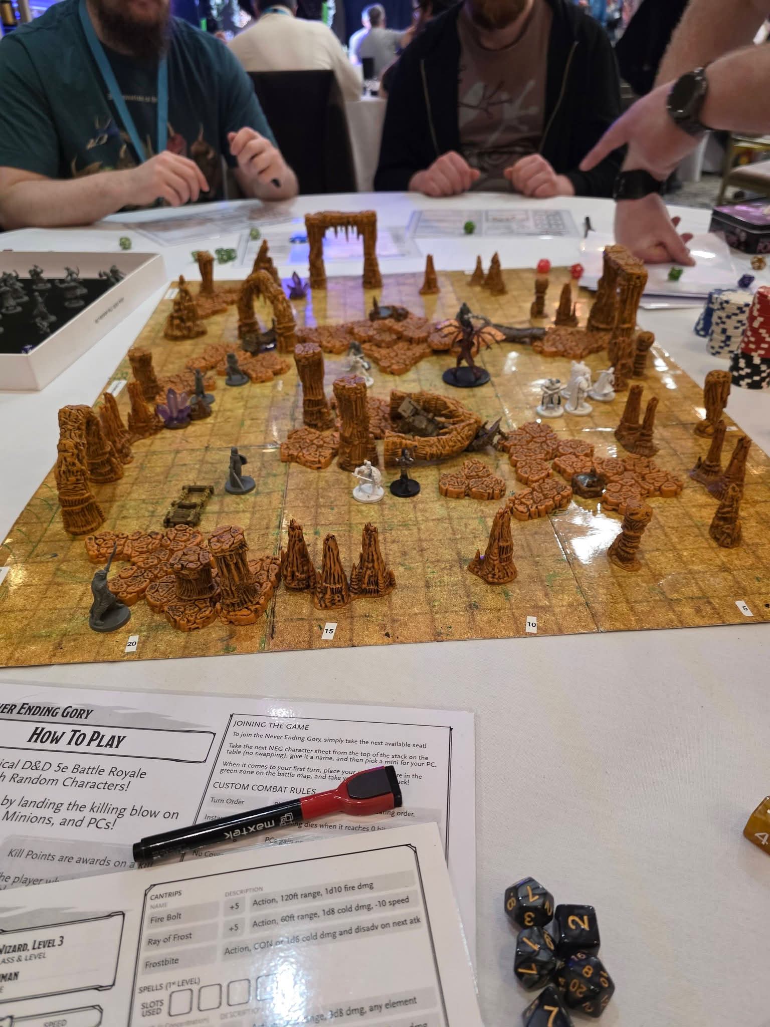 Four people play a tabletop role-playing game with a detailed 3D terrain map, dice, character sheets, and various miniatures, including pillars and archways, representing a fantasy battle scene.