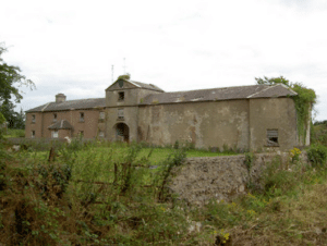 An old, weathered stone building with a large archway, surrounded by overgrown grass and bushes, under a cloudy sky.