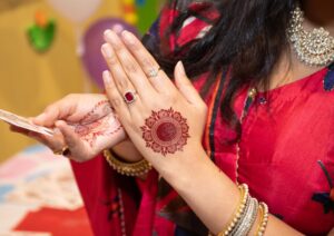 Woman in a red sari displays intricate henna designs on her hands, adorned with rings, gold bangles, and a necklace, with colourful blurred decorations in the background.