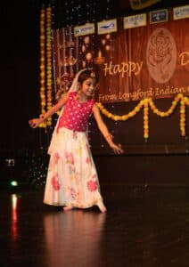 A young girl in a pink and white traditional outfit dances on stage, smiling, with festive decorations and a "Happy Diwali" banner behind her.
