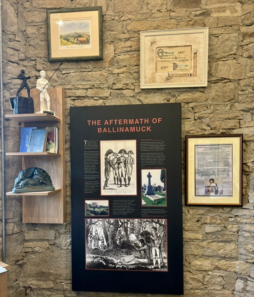 A museum display on a stone wall features "The Aftermath of Ballinamuck" panel, historic images, documents, a bust, books, and framed art, providing historical context and artefacts.
