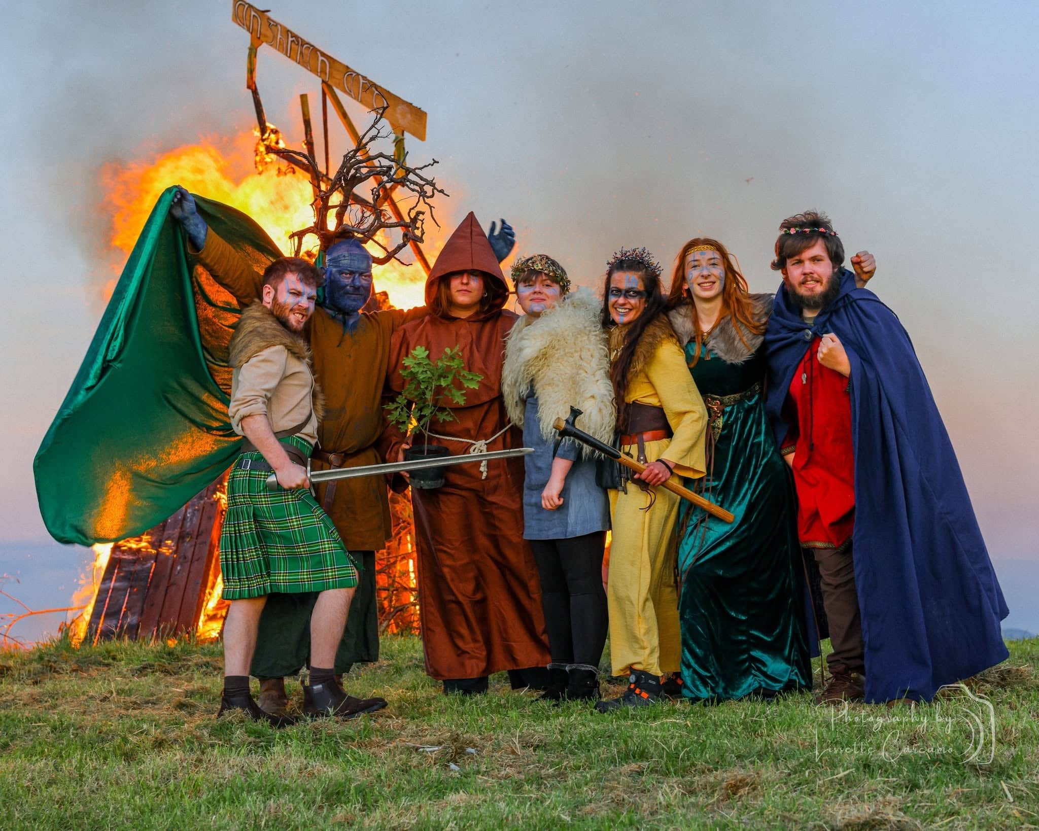 A group in fancy-dress costumes poses on grass with a burning wooden figure in the background, smiling and holding props, possibly at a festival or event.