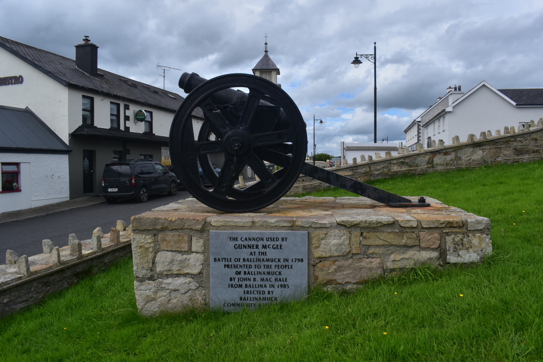 A historic cannon monument on a stone plinth with a plaque, situated on a grassy area in front of white buildings under a cloudy sky.